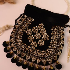 May include: A black velvet pouch with gold embroidery and beaded detailing. The pouch has a drawstring closure and black tassels with gold accents. A pearl necklace is in the background. The pouch is decorated with floral patterns.