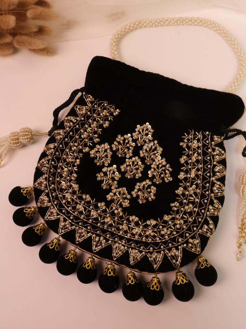 May include: A black velvet pouch with gold embroidery and beaded detailing. The pouch has a drawstring closure and black tassels with gold accents. A pearl necklace is in the background. The pouch is decorated with floral patterns.