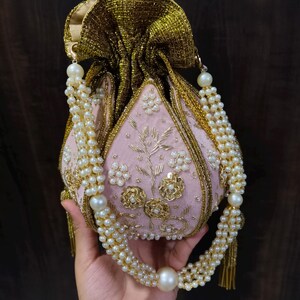 May include: A pink, beaded pouch with gold trim and a pearl-adorned handle. The pouch features floral embroidery and a gold, textured drawstring closure. The handle is made of multiple strands of pearls, with a large pearl at the center.