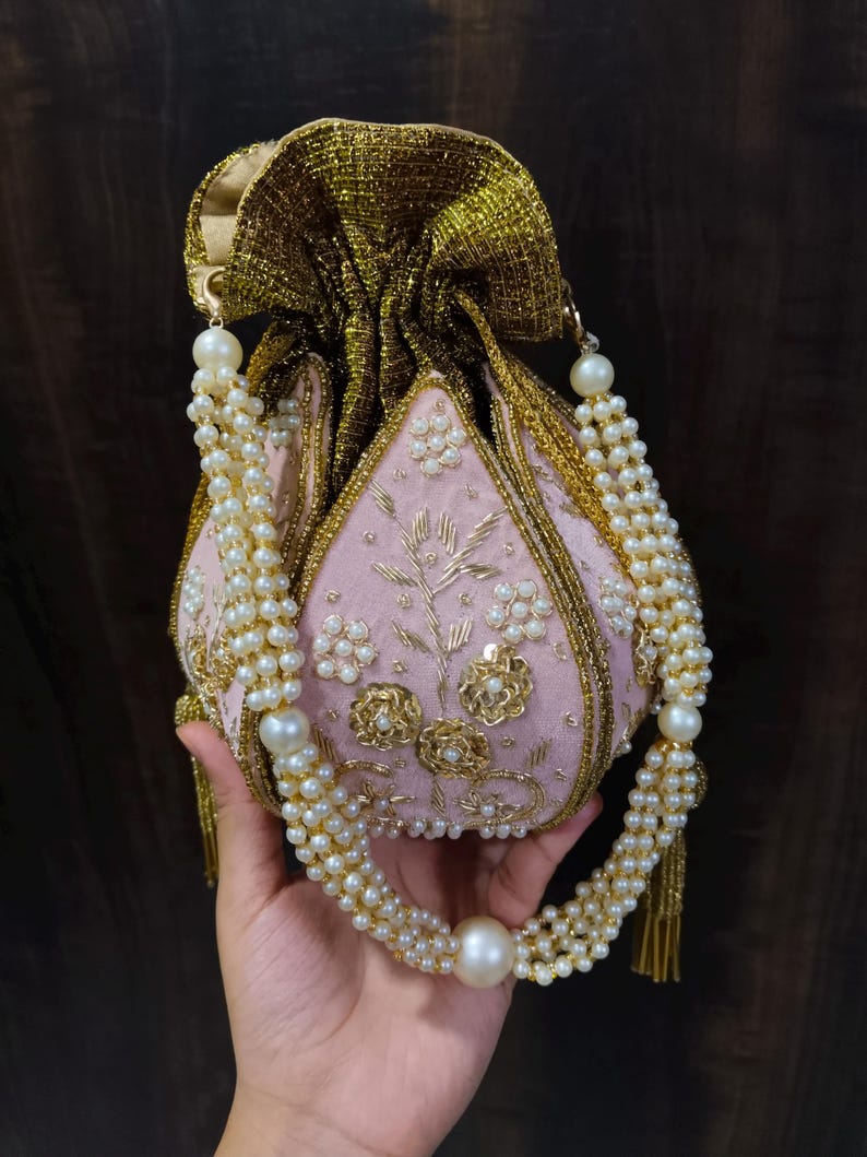 May include: A pink, beaded pouch with gold trim and a pearl-adorned handle. The pouch features floral embroidery and a gold, textured drawstring closure. The handle is made of multiple strands of pearls, with a large pearl at the center.