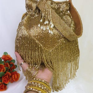 May include: A gold beaded drawstring bag with a matching strap and fringe. The bag is embellished with small white pearl-like beads. The bag is held in a hand, with yellow bangles on the wrist.