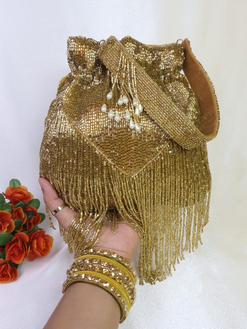 May include: A gold beaded drawstring bag with a matching strap and fringe. The bag is embellished with small white pearl-like beads. The bag is held in a hand, with yellow bangles on the wrist.