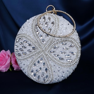 Puede incluir: Un bolso de mano redondo blanco con cuentas y un asa dorada. El bolso de mano está adornado con brillantes pedrería transparente y pequeñas cuentas blancas. El diseño presenta un patrón floral. El fondo es una tela azul oscuro con rosas rosas.