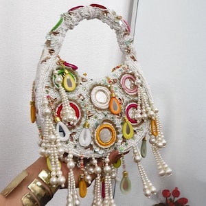 Könnte beinhalten: Eine halbmondförmige Handtasche, die mit weißen Perlen bedeckt ist und kreisförmige Stickereiaufnäher mit zentralen Spiegeln, farbige Akzente in Gelb, Pink und Grün sowie herabhängende Stränge aus weißen Kunstperlen und tropfenförmigen Anhängern aufweist.