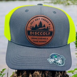 Könnte beinhalten: Graue und neongelbe Trucker-Kappe mit einem braunen Leder-Patch, auf dem "Get Out There & Disc Golf, Bang Chains & Take Names" steht. Der Patch zeigt eine Silhouette von Bäumen und einem Disc-Golf-Korb. Die Kappe hat außerdem einen kleinen schwarzen und silbernen Aufkleber mit der Aufschrift "BK Caps Since 2013".