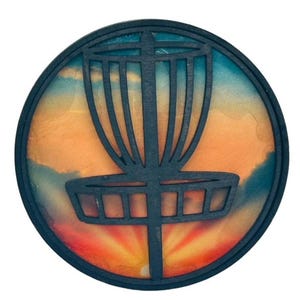 Puede incluir: Un disco de madera con una silueta negra de una cesta de disco golf frente a un fondo de puesta de sol.