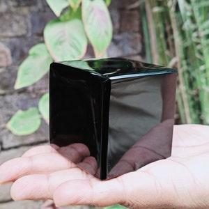Op de afbeelding: Een zwarte obsidiaan kubus, een soort natuurlijk glas, wordt in de hand van een persoon gehouden. De kubus is glad en gepolijst en reflecteert het licht.