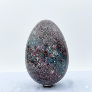 Puede incluir: Una escultura de piedra en forma de huevo con una superficie moteada. La piedra presenta una mezcla de colores, incluyendo morado oscuro, verde azulado y gris. La escultura tiene un aspecto pulido y se asienta sobre una pequeña base plateada.