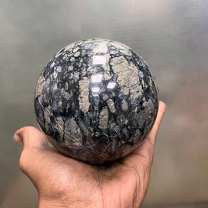Könnte beinhalten: Eine polierte Kugel aus schwarzem und grauem Stein mit einem wirbelnden Muster aus weißen und blauen Einschlüssen.