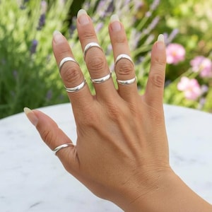 May include: A set of four silver-colored rings worn on the fingers. Three of the rings feature a distinctive open-arch design that sits across the knuckle, while the fourth ring is a simple, thin band worn on the thumb. The metal has a smooth finish.