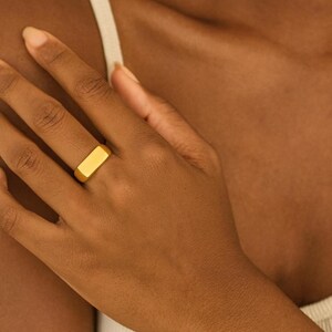 Puede incluir: Un anillo dorado con una cara rectangular se lleva en un dedo. El anillo tiene una superficie lisa y pulida y un diseño simple y moderno. El anillo es una joya.