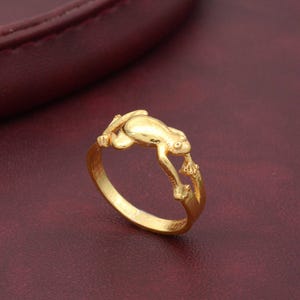 Könnte beinhalten: Ein goldfarbener Ring mit einem detaillierten Froschdesign. Der Frosch ist auf dem Ringband platziert, seine Beine erstrecken sich um das Band.