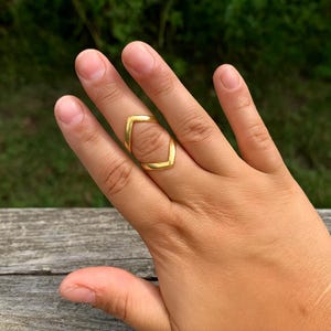May include: A gold-colored ring with a geometric design. The ring features a square shape with rounded corners, creating a modern and minimalist aesthetic. The ring is worn on a finger.