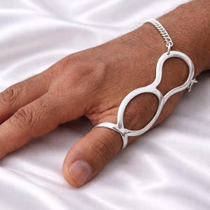 May include: A silver-toned hand chain bracelet featuring a ring for the thumb and a chain connecting to a double-lobed design that rests on the back of the hand. The jewelry is displayed against a white satin background.