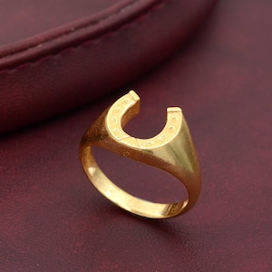 Puede incluir: Un anillo de oro con un diseño de herradura. La herradura está en relieve y tiene una superficie texturizada.