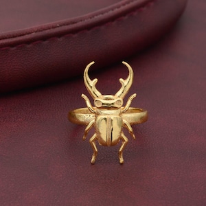 May include: A gold-coloured ring featuring a detailed beetle design. The beetle has a raised, textured body and long, pointed antennae.