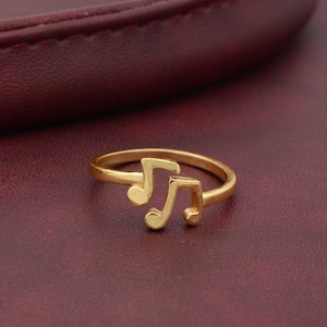 May include: A gold-colored ring with two musical note designs. The ring is on a burgundy surface.