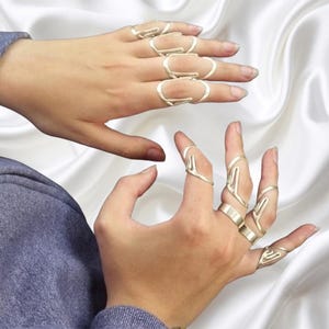 Puede incluir: Una colección de anillos de metal plateado usados en varios dedos. Los anillos presentan diseños alargados de marco abierto que se extienden a lo largo de los dedos, con algunos anillos que muestran un motivo en forma de mano con acabado pulido.