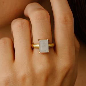May include: A gold ring featuring a rectangular, light-colored stone. The ring has a simple, elegant design with a smooth, matte finish. The stone is set in a gold bezel, creating a classic and timeless look.