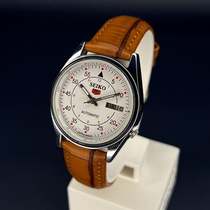 May include: A silver-toned Seiko automatic watch with a white face, featuring black numerals and red accents. The watch has a brown leather band and displays the day of the week. The watch is on a white stand.