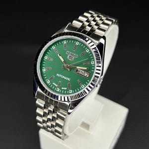 Puede incluir: Un reloj automático Seiko 5 de acero inoxidable con esfera verde. El reloj tiene una pantalla de día y fecha a las 3 en punto y una pulsera plateada. El reloj se muestra sobre un soporte blanco sobre un fondo oscuro.