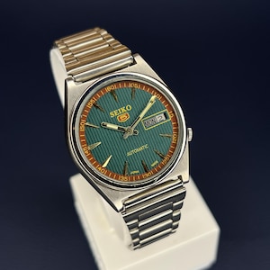 May include: A vintage Seiko 5 automatic watch with a silver-tone stainless steel band. The watch face is green with gold-tone hands and markers, and a day-date display. The watch is displayed on a white stand against a dark blue background.