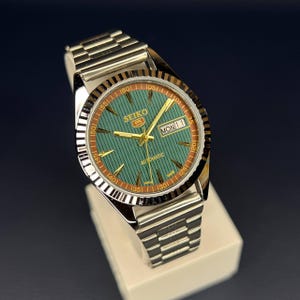 May include: A Seiko 5 automatic wristwatch with a silver-tone stainless steel band. The watch face is green with gold-tone hands and markers, and a day-date display. The bezel is brown and silver, and the watch is displayed on a white surface.