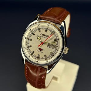 May include: A silver-toned Citizen automatic watch with a brown leather band. The watch face displays the day and date, with red and black accents. The watch is displayed on a white stand.