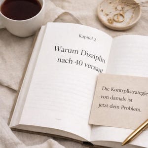 May include: An open book with German text, a sticky note, and a pen. The book's title reads "Warum Disziplin nach 40 versagt." A cup of coffee and a small dish with jewelry are also present.