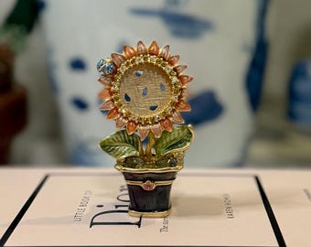 May include: A small, gold-toned, decorative sunflower trinket box with a blue and white patterned interior. The box is sitting on top of two books, one titled "Little Book of Dior" and the other titled "Think Like a Decorator".