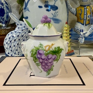 May include: A white porcelain sugar bowl with a grape design. The bowl has a lid topped with a purple grape cluster and a small spoon. The design features green leaves and purple grapes. The bowl is sitting on a book.