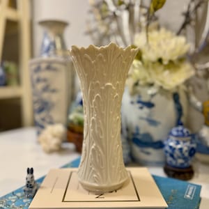 May include: A white porcelain vase with an intricate floral design. The vase has a scalloped rim and a tall, slender shape.