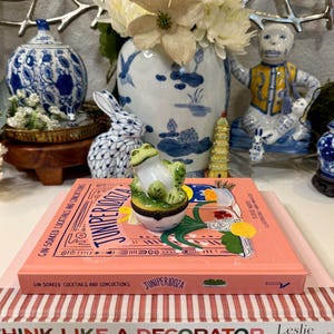 May include: A decorative arrangement featuring a frog figurine, a book titled "Juniperiooza", and various porcelain pieces. The frog is green and white. The book is pink with white and red striped book below it. A blue and white vase holds white flowers.