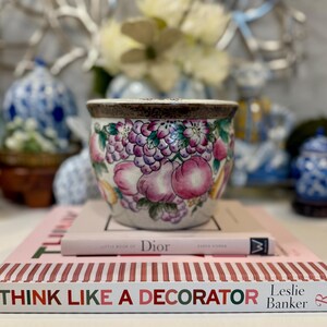 May include: A decorative ceramic bowl with a fruit design in shades of pink, purple, and green. The bowl sits atop two books, one titled "Dior" and the other "Think Like a Decorator."