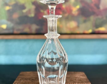May include: A clear glass decanter with a faceted design and a matching stopper. The decanter has a teardrop shape and sits on a wooden surface. The stopper is a geometric shape. The background is a colorful abstract painting.