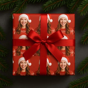 May include: A wrapped gift with red wrapping paper featuring a repeating pattern of a smiling person wearing a Santa hat and a red sweater. A red satin ribbon and bow are tied around the gift. Green pine branches are in the background.