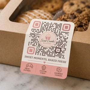 May include: A cardboard bakery box with a clear window, filled with cookies. A pink and white label with a QR code and the text "Sweet Crumbs Bakery" is attached to the box. The label also says "Sweet Moments, Baked Fresh."