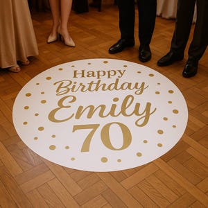 Op de afbeelding: Een witte, ronde vloersticker met gouden tekst "Happy Birthday Emily 70". De sticker is versierd met gouden stippen en is geplaatst op een houten vloer. Er staan mensen rond de sticker.