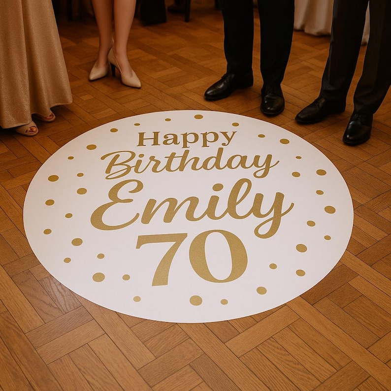 Op de afbeelding: Een witte, ronde vloersticker met gouden tekst "Happy Birthday Emily 70". De sticker is versierd met gouden stippen en is geplaatst op een houten vloer. Er staan mensen rond de sticker.
