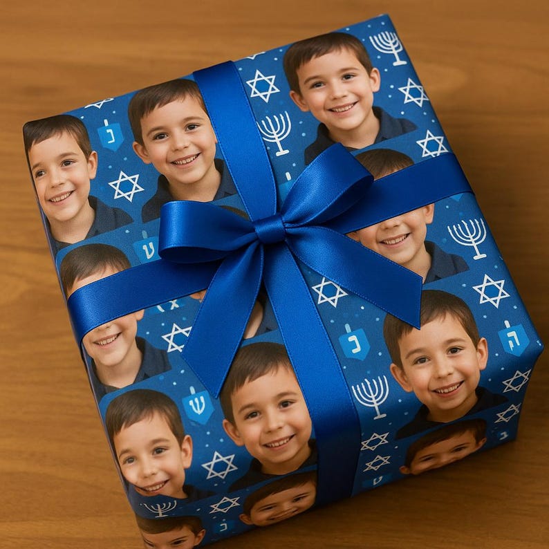 May include: A wrapped gift with blue wrapping paper featuring a pattern of a smiling child's face, Stars of David, menorahs, and dreidels. A blue satin ribbon and bow secure the gift.