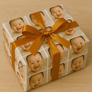 May include: A wrapped gift box featuring a repeating pattern of a smiling baby's face. The wrapping paper is tan with a grid design. A gold-colored ribbon is tied in a bow on top of the box, adding a decorative touch.