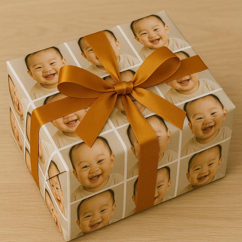 May include: A wrapped gift box featuring a repeating pattern of a smiling baby's face. The wrapping paper is tan with a grid design. A gold-colored ribbon is tied in a bow on top of the box, adding a decorative touch.