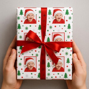 May include: A wrapped gift with a red ribbon and bow. The wrapping paper is white with a pattern of red and green Christmas trees, red dots, and photos of a baby wearing a Santa hat.