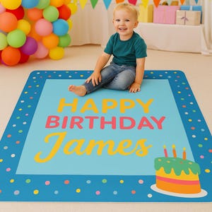 Op de afbeelding: Een blauwe verjaardagsmat met de woorden "HAPPY BIRTHDAY James" in geel en roze. Een kleurrijke taartafbeelding staat in de rechter benedenhoek. Een jong kind zit op de mat.