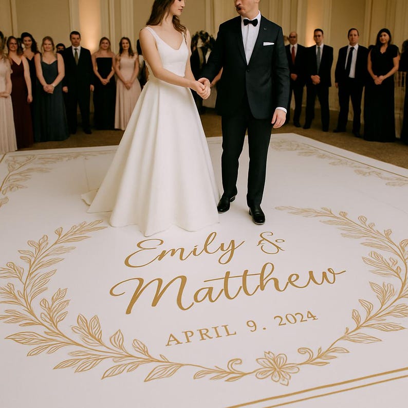 Op de afbeelding: Een witte trouwdansvloer met de namen "Emily & Matthew" en de datum "April 9, 2024" in gouden letters. De vloer heeft een gouden bloemenrand en is omringd door bruiloftgasten.