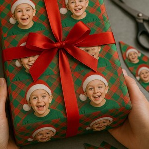 May include: A wrapped gift featuring a repeating pattern of a smiling child wearing a Santa hat and green shirt. The wrapping paper has a red and green plaid design. A large red ribbon is tied around the gift. Scissors are nearby.