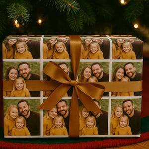 May include: A wrapped gift with a custom photo collage on the wrapping paper. The gift is tied with a wide, golden-brown ribbon and bow. The gift is under a Christmas tree with a red ornament.