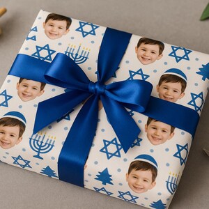 May include: A wrapped gift with a blue satin bow. The wrapping paper is white with a pattern of smiling faces, Stars of David, menorahs, and blue trees. A wooden dreidel sits nearby.