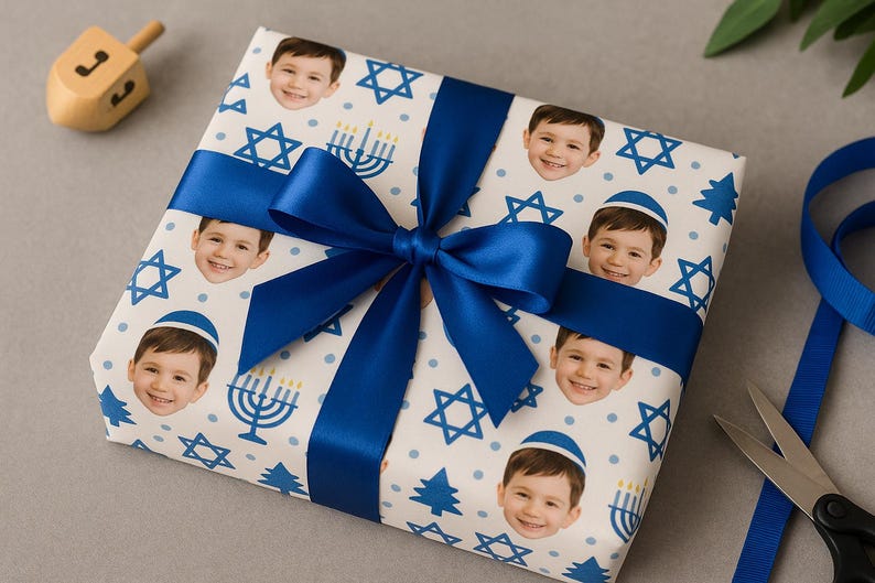 May include: A wrapped gift with a blue satin bow. The wrapping paper is white with a pattern of smiling faces, Stars of David, menorahs, and blue trees. A wooden dreidel sits nearby.