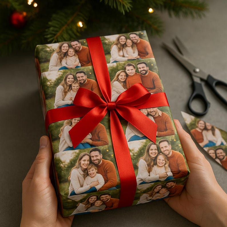May include: A wrapped gift featuring a family photo pattern, tied with a vibrant red satin ribbon and bow. The gift is held by hands, with scissors and a piece of the wrapping paper visible in the background. The gift is ready for the holidays.